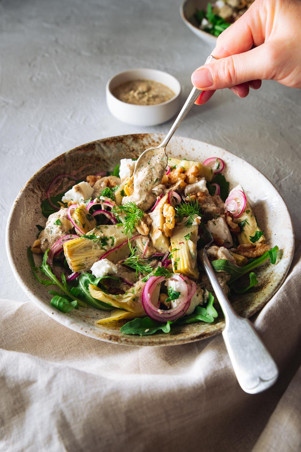 A hand drizzling yoghurt dressing over an artichoke salad in a salad bowl.