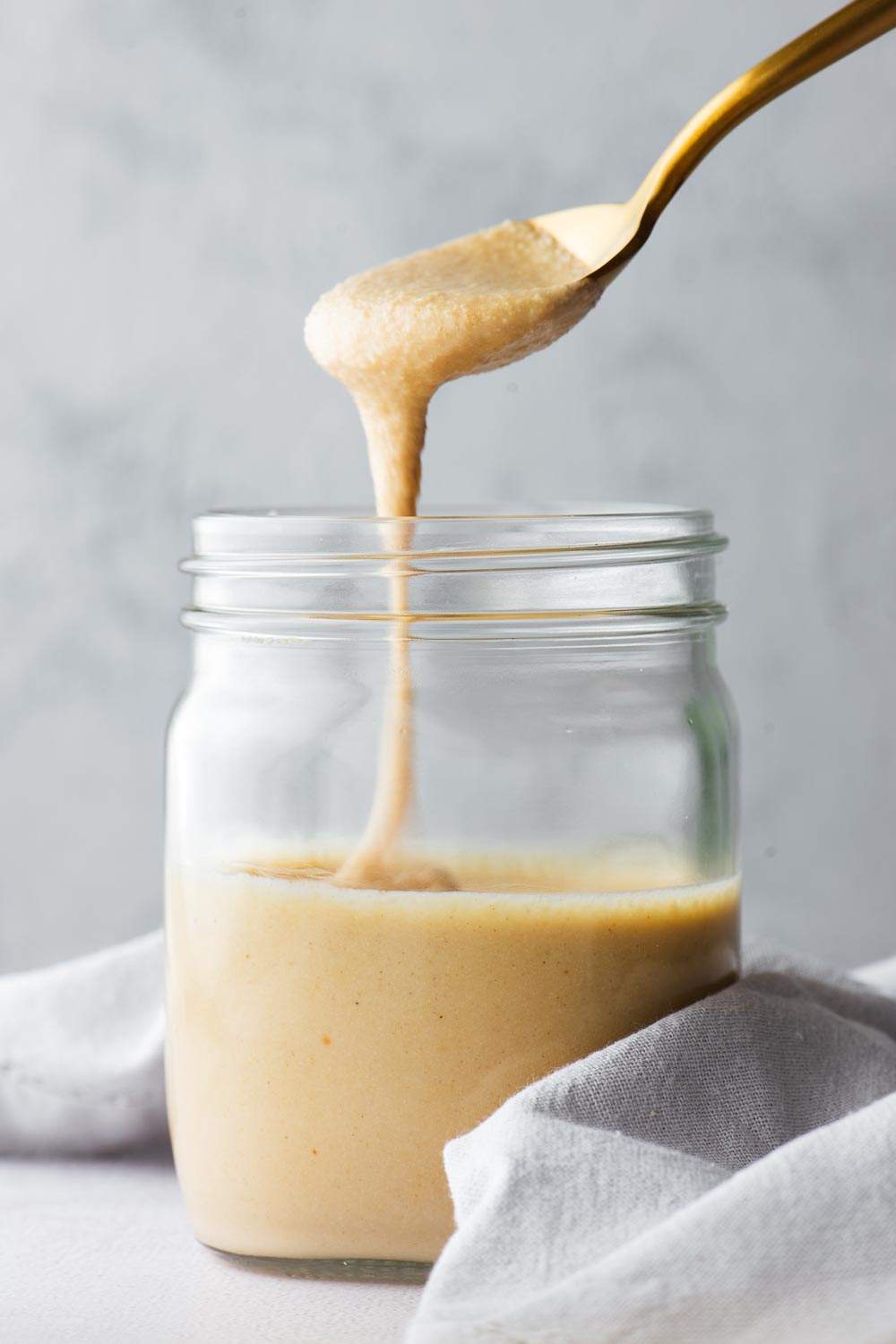 Thick but pourable homemade tahini drizzled from a gold spoon into a half-full mason jar.