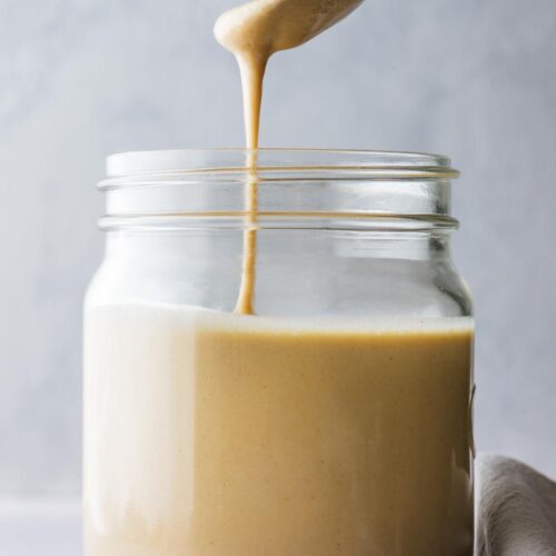 A side angle view of a spoon drizzling homemade tahini into a mason jar against a grey background.