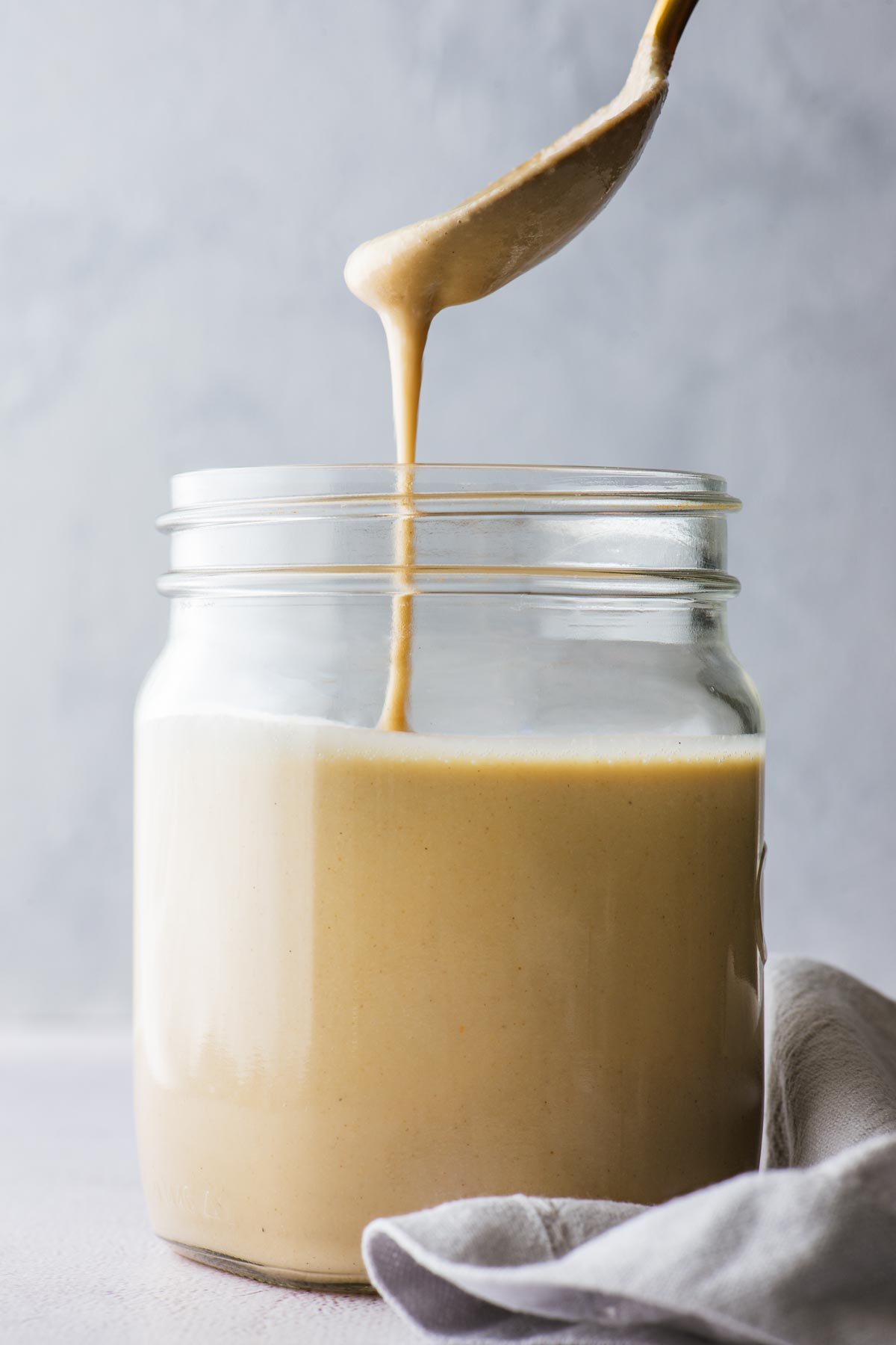 A side angle view of a spoon drizzling homemade tahini into a mason jar against a grey background.
