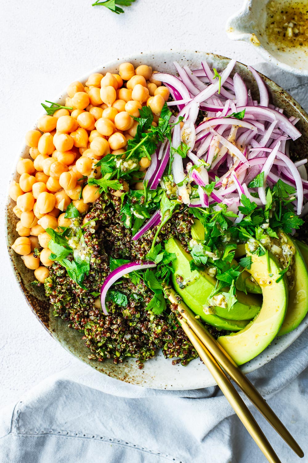A kale and quinoa salad with chickpeas, avocado, red onion, and preserved lemon dressing in a ceramic bowl with gold cutlery.