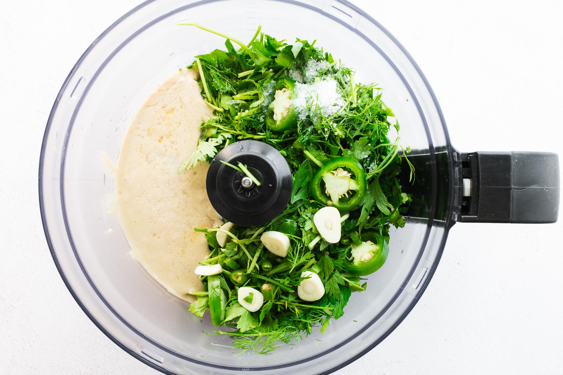Tahini, fresh herbs, raw garlic and chillies in a food processor as viewed from above.