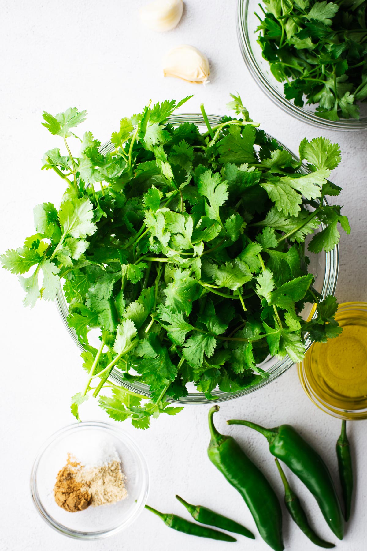 Coriander leaves in a glass bowl viewed from above, arranged in a flatlay with chillies, spices, olive oil, parsley and garlic cloves.
