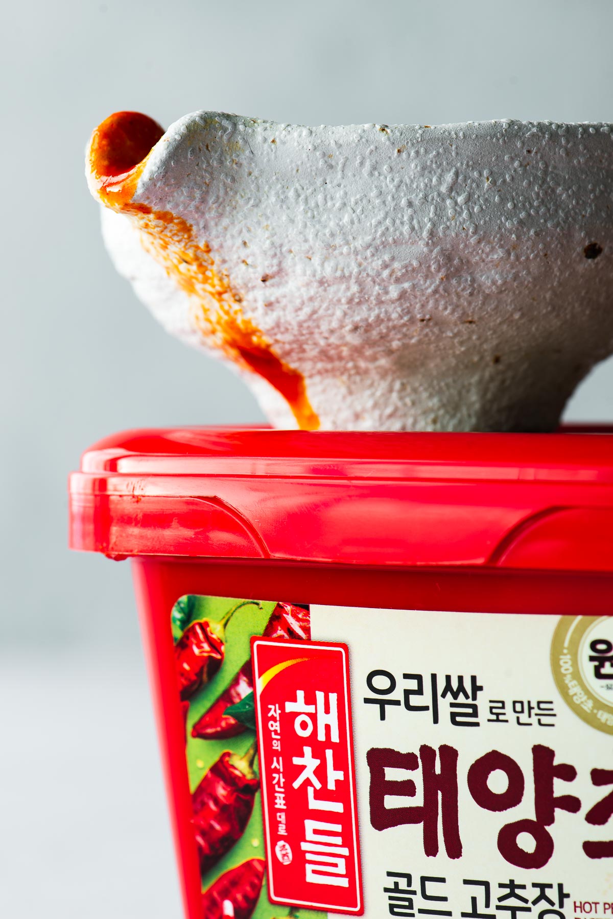 Side angle view of a pourer with red bibimbap sauce drizzling from the spout, on top of a red tub of gochujang paste.