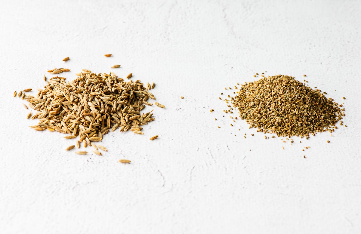 A small pile of cumin seeds next to a pile of celery seeds.