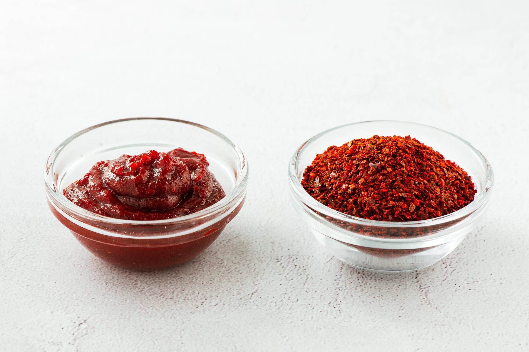 Gochugaru chilli flakes and gochujang paste in two small glass bowls on a concrete surface.