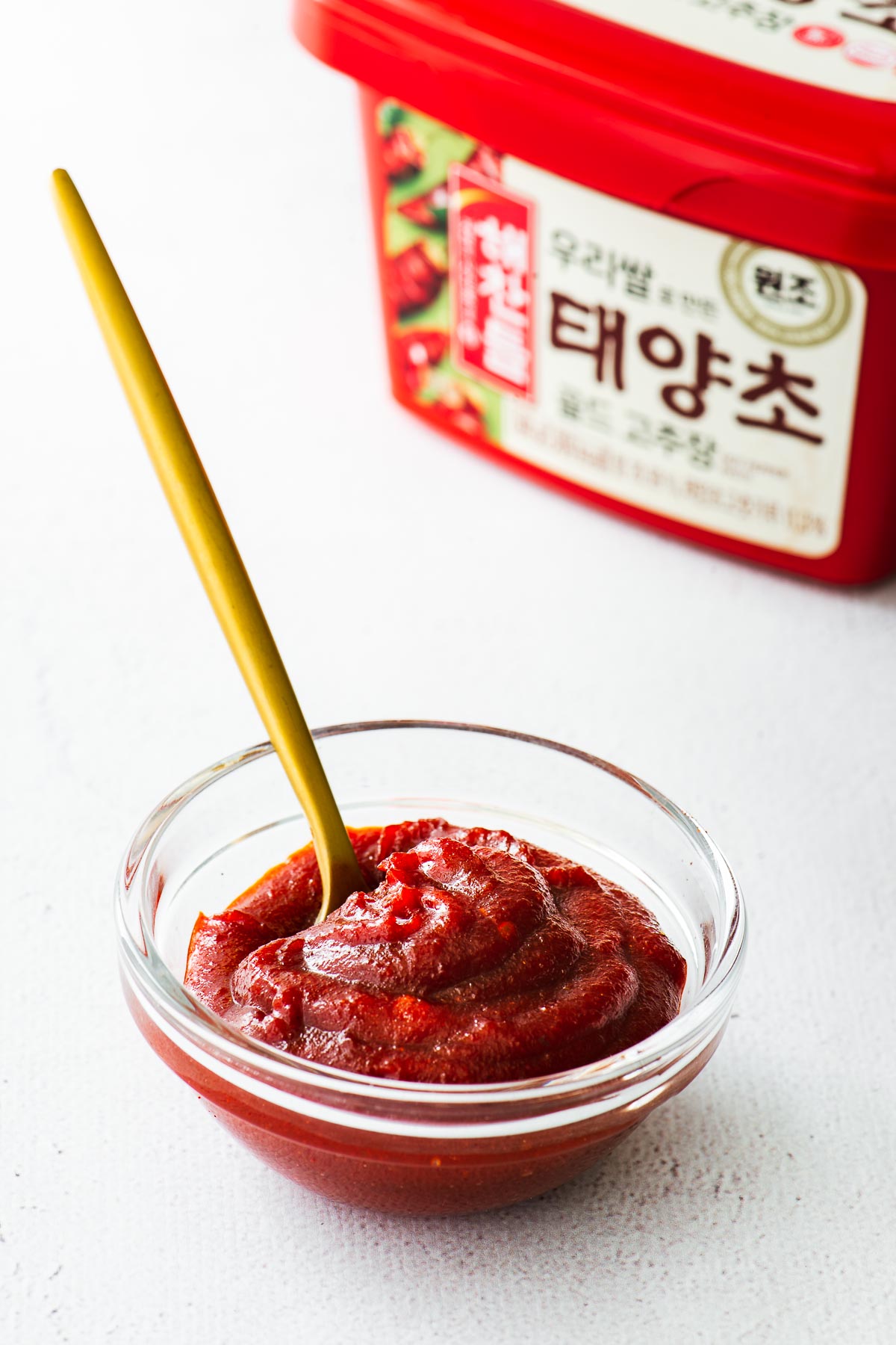 A glass bowl with gochujang paste and a gold spoon with the classic red gochujang tub in the background.