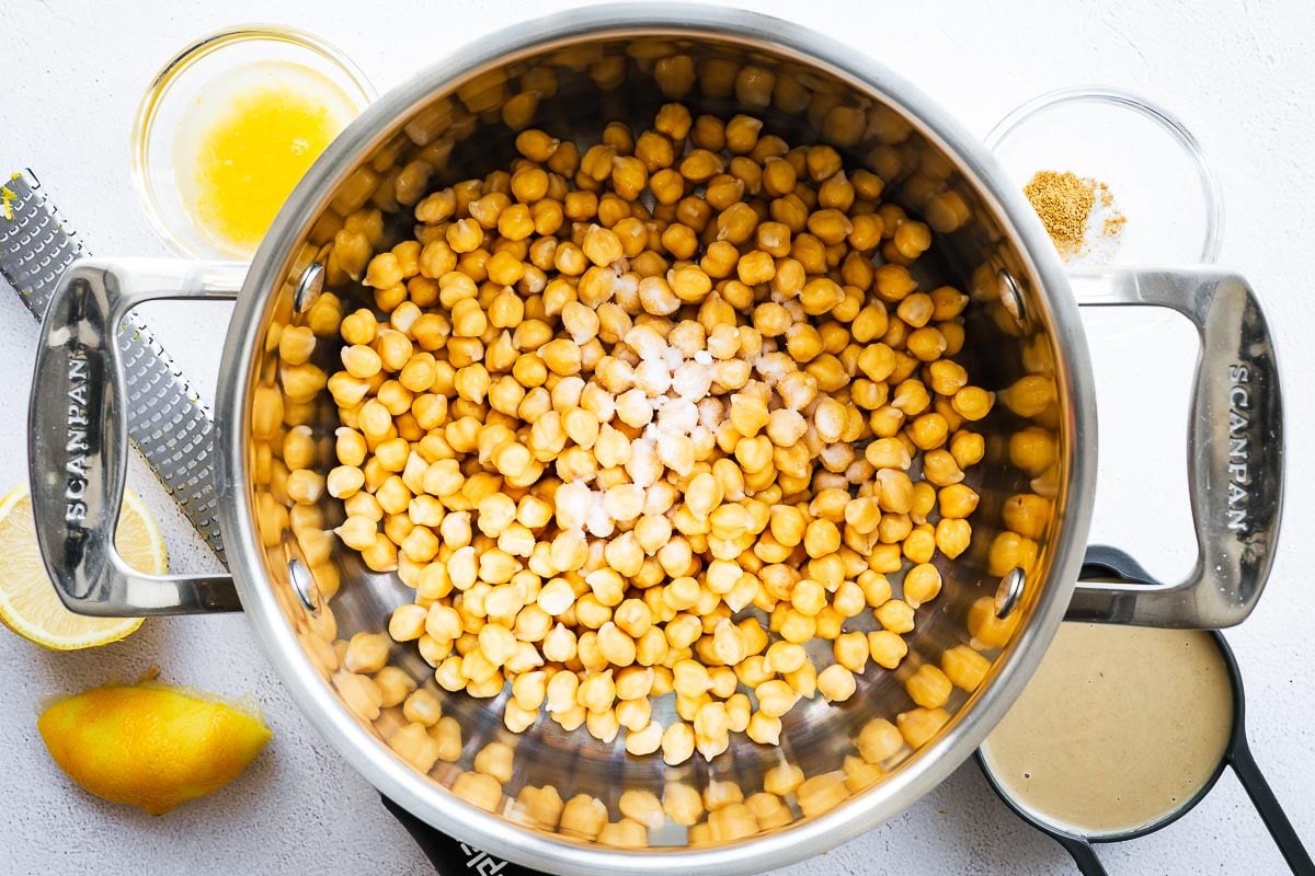 Top down view of dried chickpeas that were soaked overnight in a stainless steel pot surrounded by hummus ingredients.