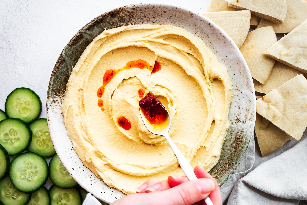 Top down view of a bowl of hummus with a hand drizzling harissa oil over the smooth top.