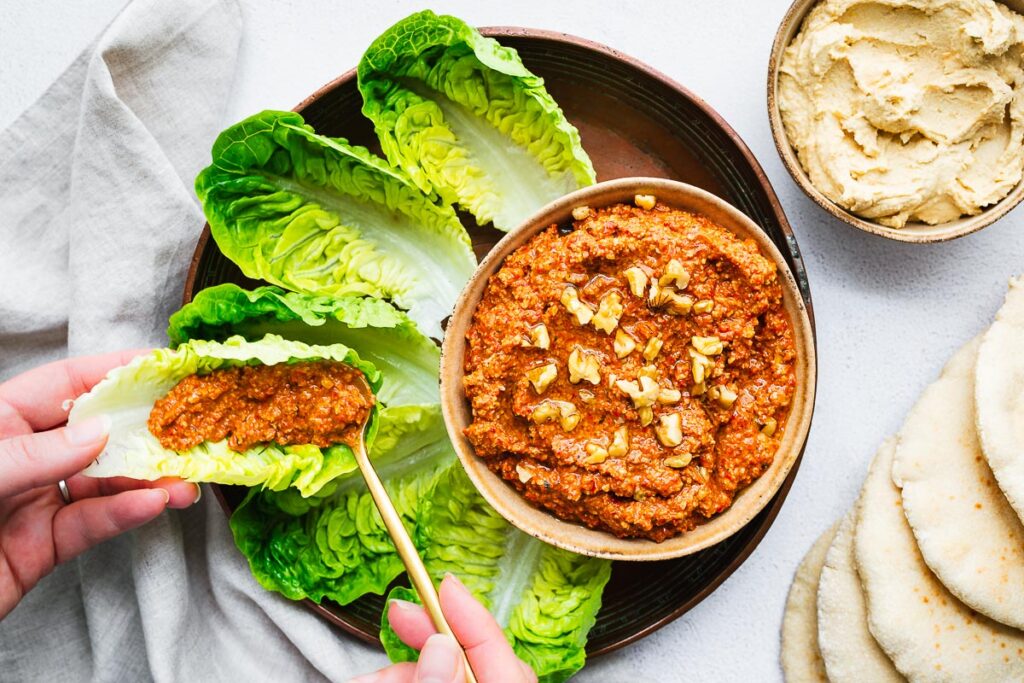 Healthy red pepper and walnut dip, called muhammara, served as a healthy snack with lettuce cups. Top down view with a hand scooping the red pepper dip with a golden spoon.