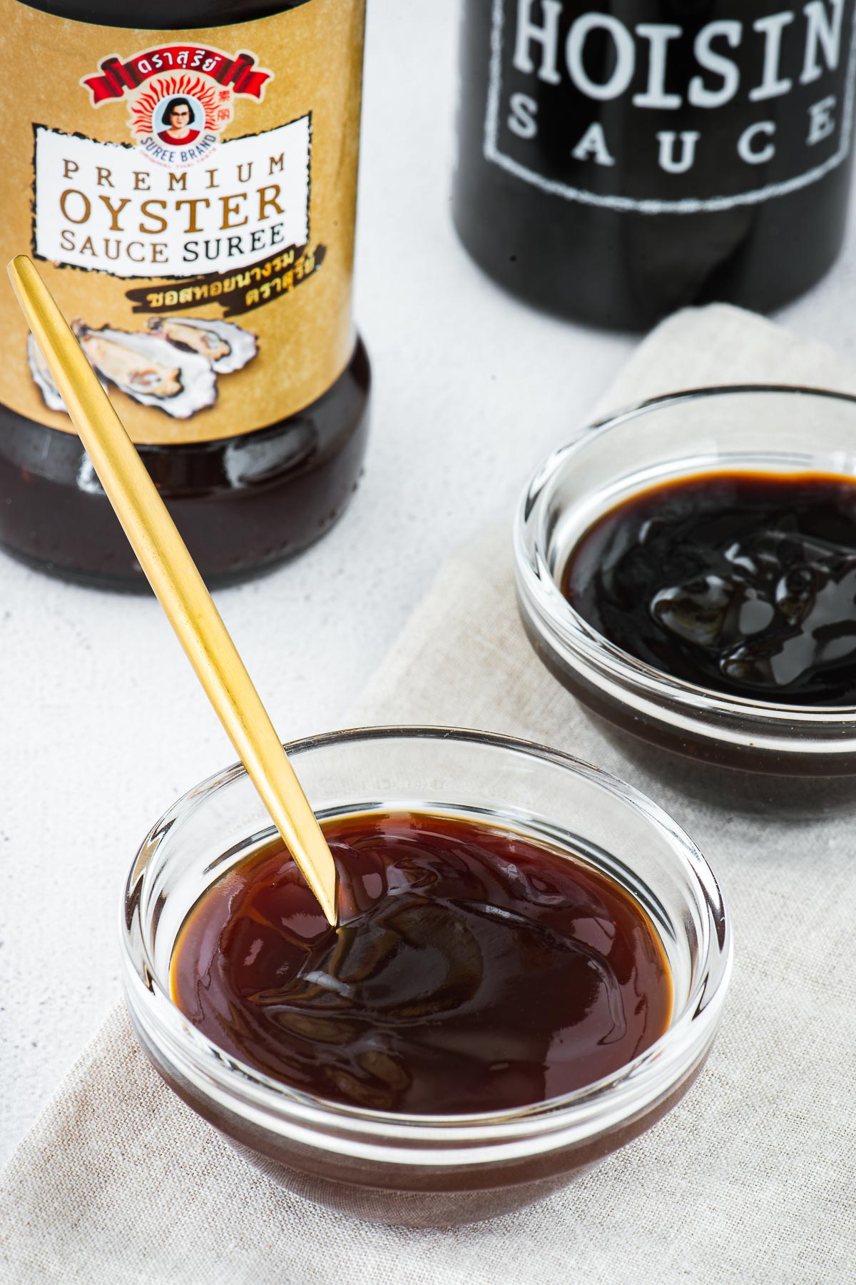 Hoisin sauce vs oyster sauce in small glass bowls with bottles in the background.