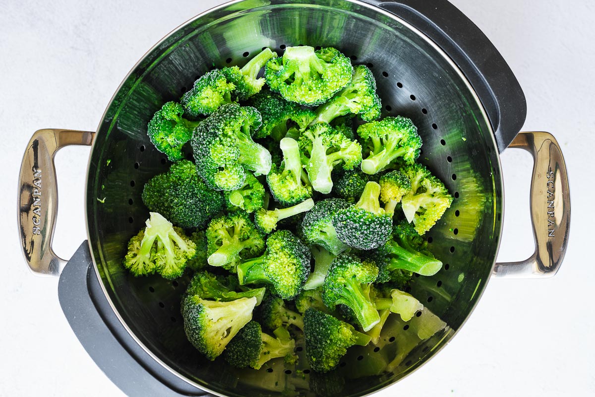 Frozen broccoli florets in a colander set over a stainless steel pot ready for steaming.