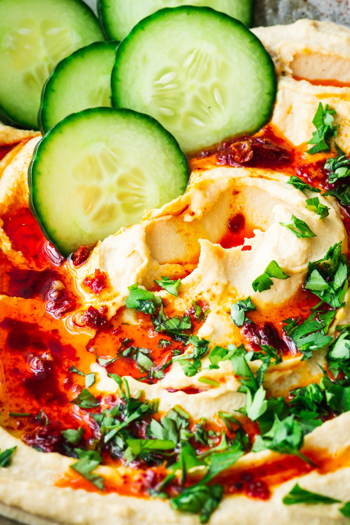 A close-up of fluffy hummus drizzled with harissa oil and served with cucumber slices.