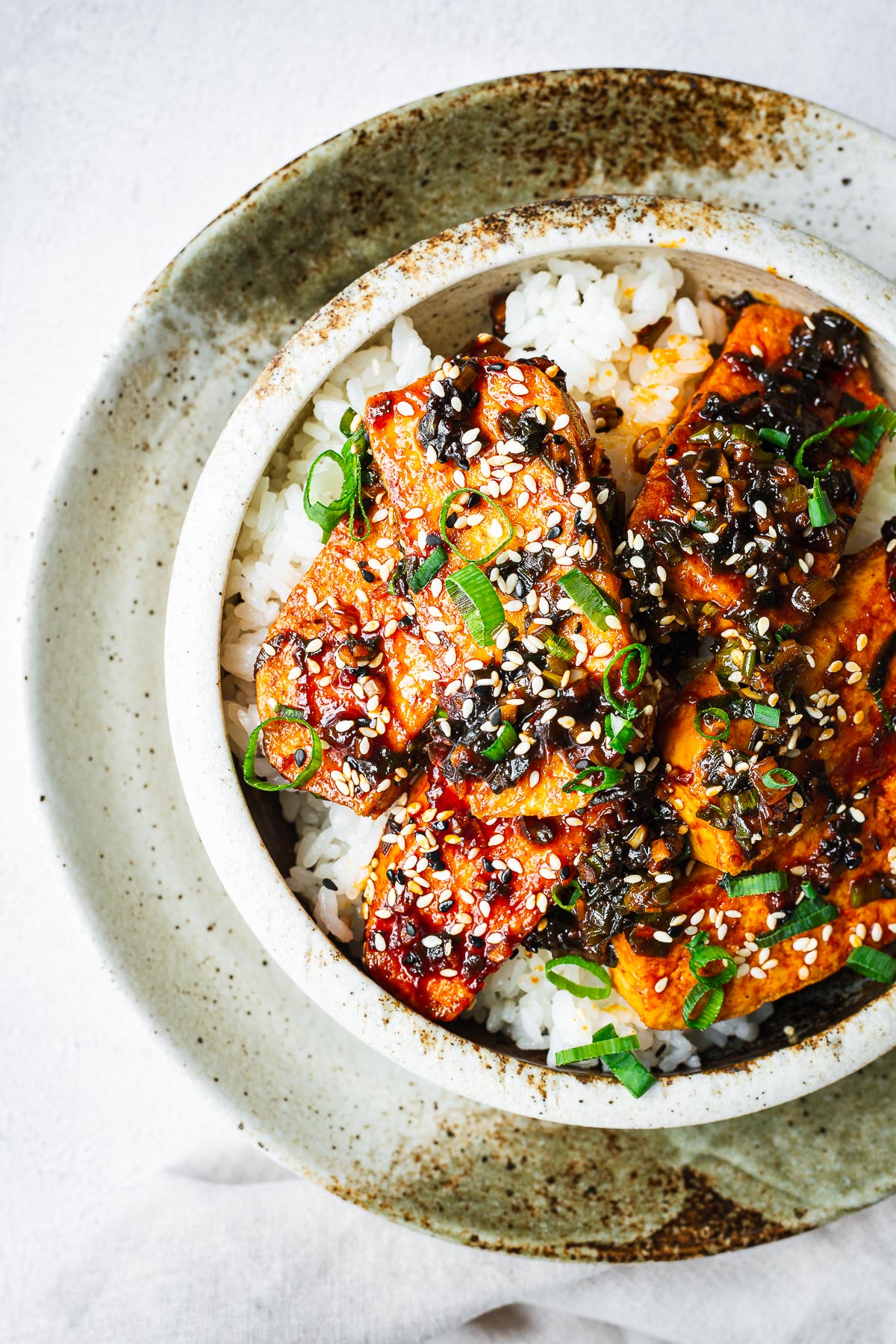 Top down close-up of a ceramic bowl with white rice topped with Korean braised tofu, sesame seeds and green onion.