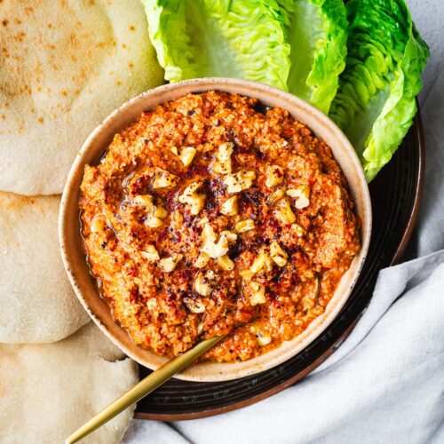 Top down view of a bowl of Lebanese muhammara surrounded by lettuce and fresh pita bread.