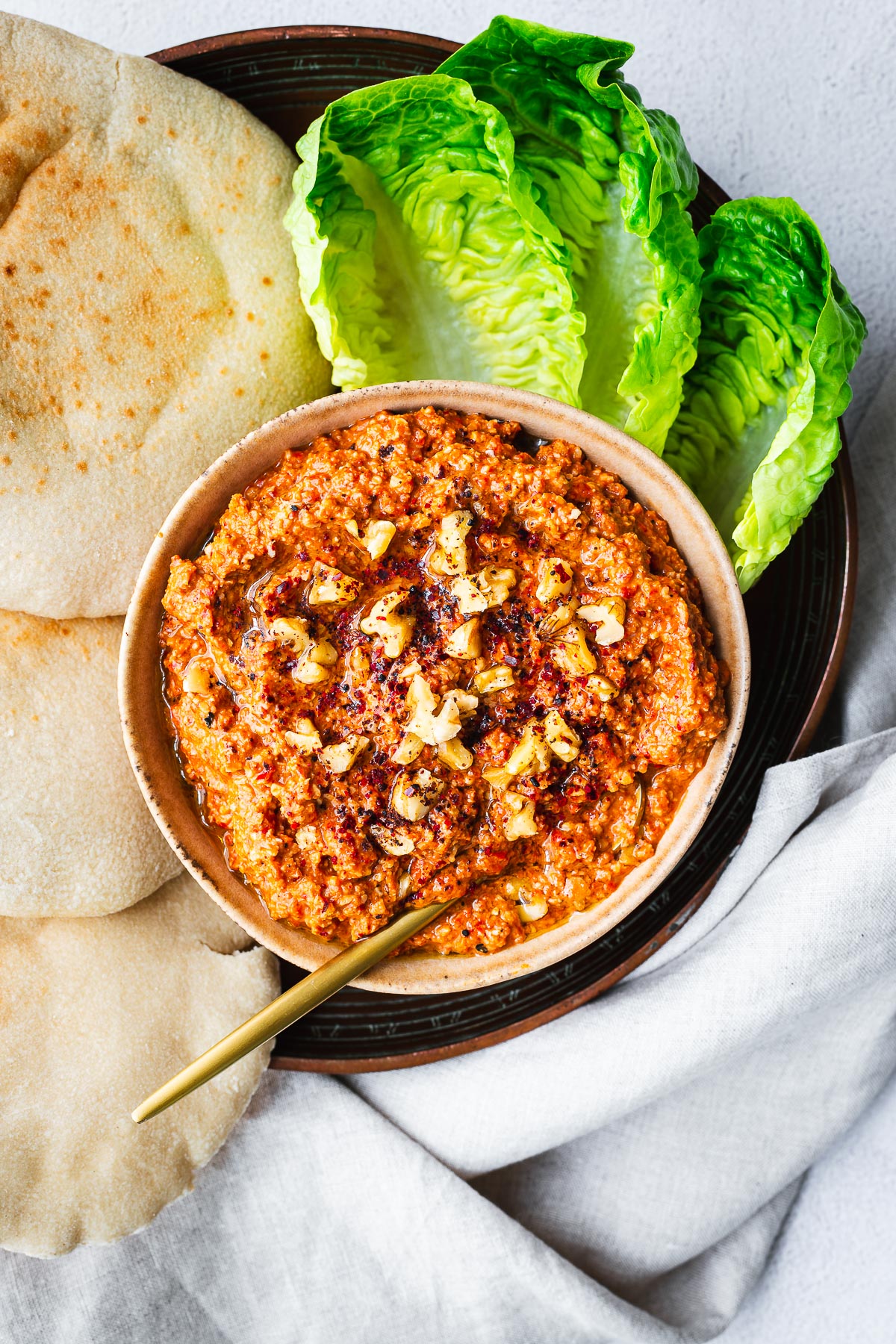 Top down view of a bowl of Lebanese muhammara surrounded by lettuce and fresh pita bread.