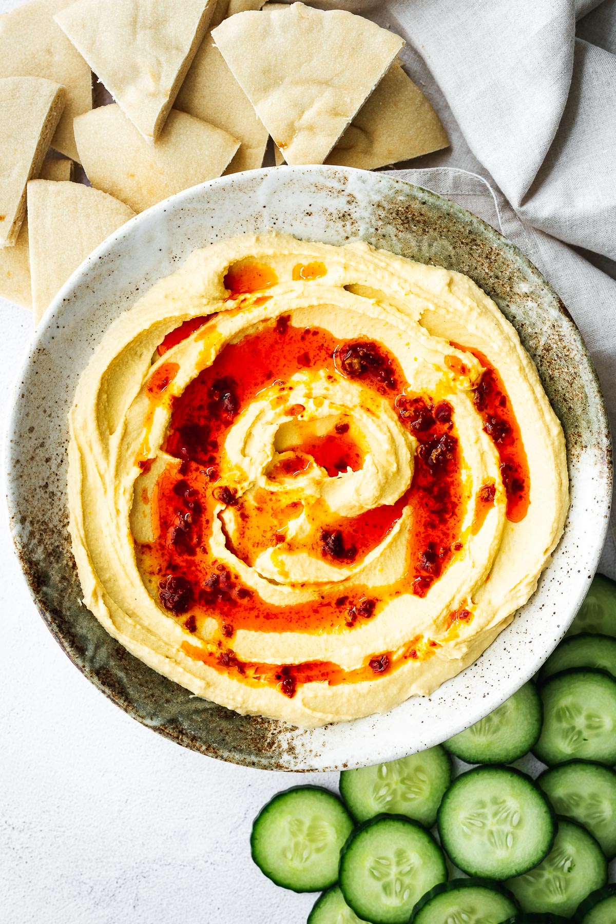 Top down view of a bowl of lemon hummus without garlic drizzled with harissa oil and served with cucumber slices and pita triangles.
