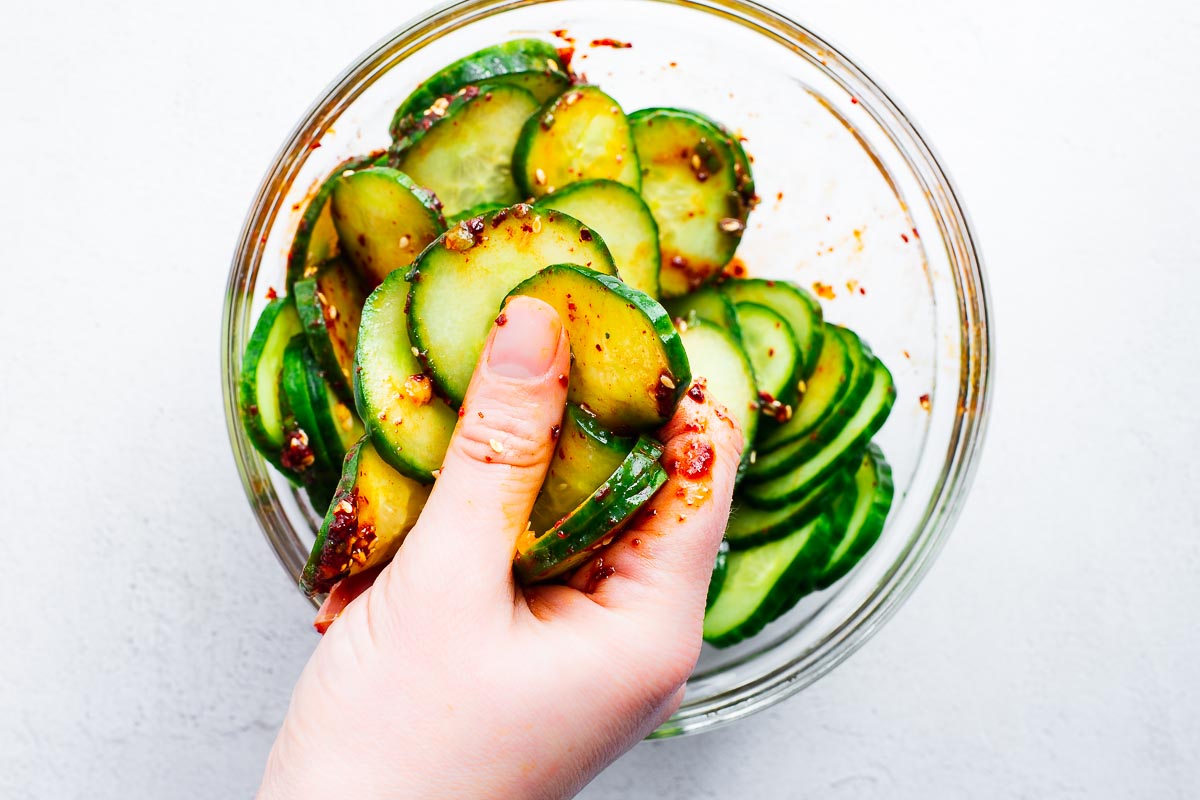 A hand mixing cucumbers with spicy Korean gochugaru dressing in a glass bowl.