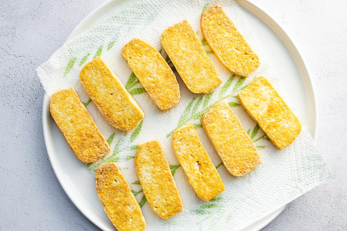 Top down view of crispy pan-fried tofu slices arranged on a white plate with kitchen towel.