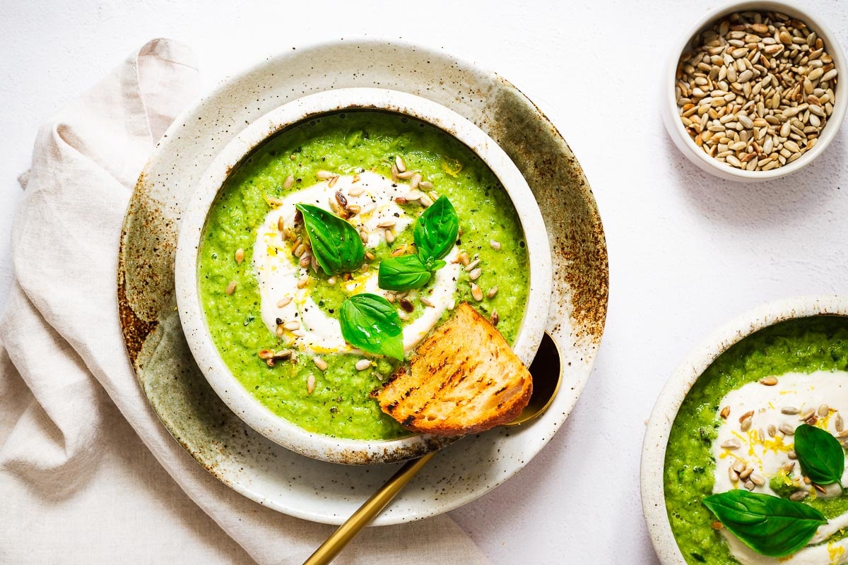 Ceramic bowls with plant-based broccoli soup topped with cashew cream, sunflower seeds and fresh basil leaves.