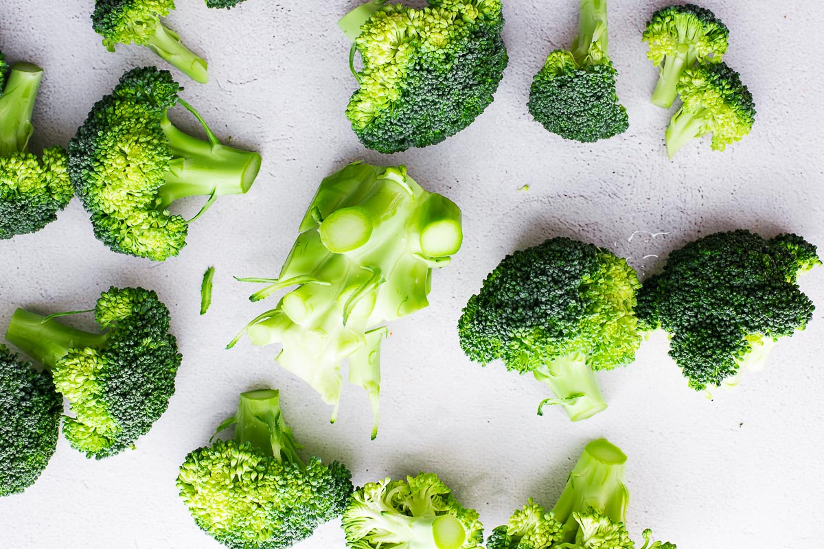 Broccoli florets and a unprocessed broccoli stalk viewed from above, arranged on a cement background.