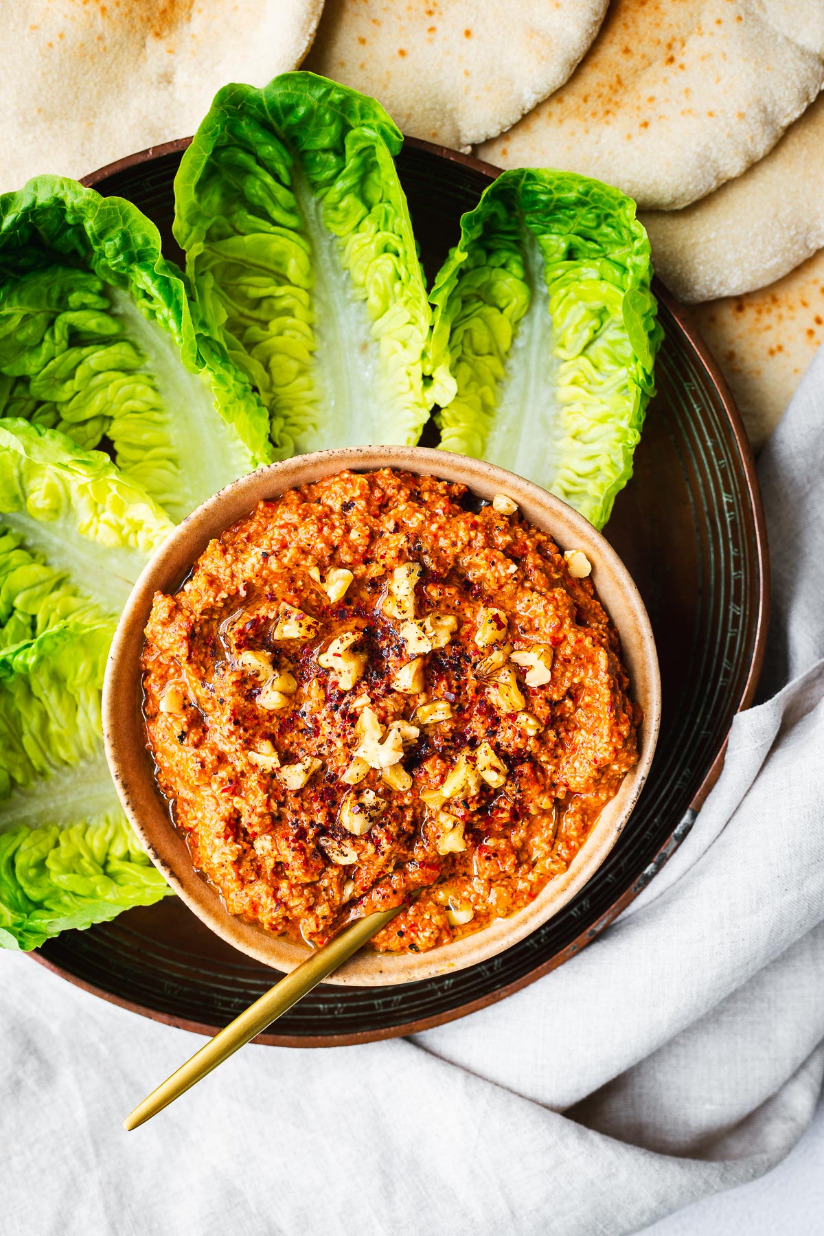 Top down view of a bowl with red pepper and walnut dip surround by lettuce leaves and fresh pita bread.