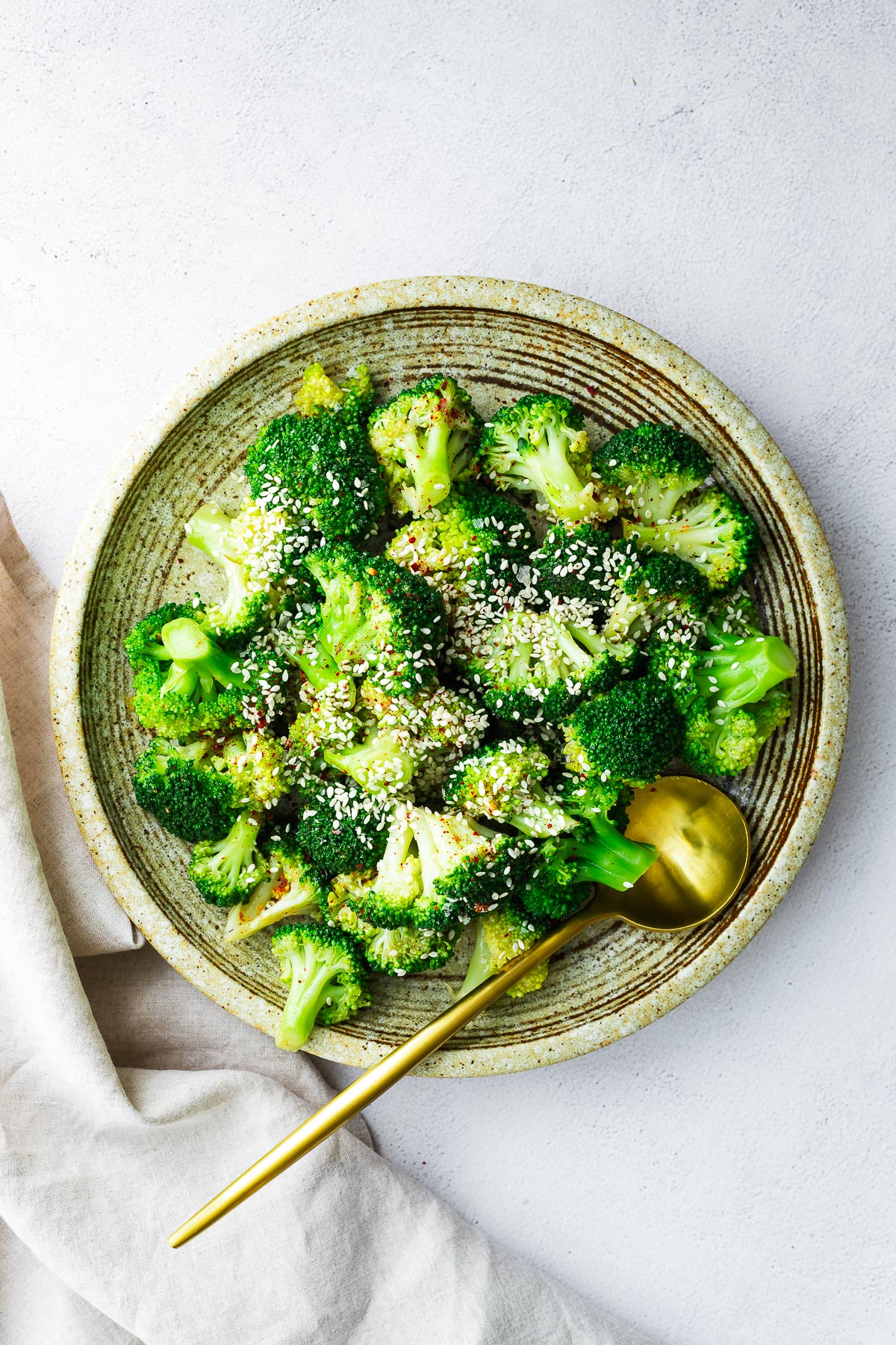 Steamed frozen broccoli with a sweet Asian dressing sprinkled with sesame seeds, served on a ceramic plate with a gold spoon.