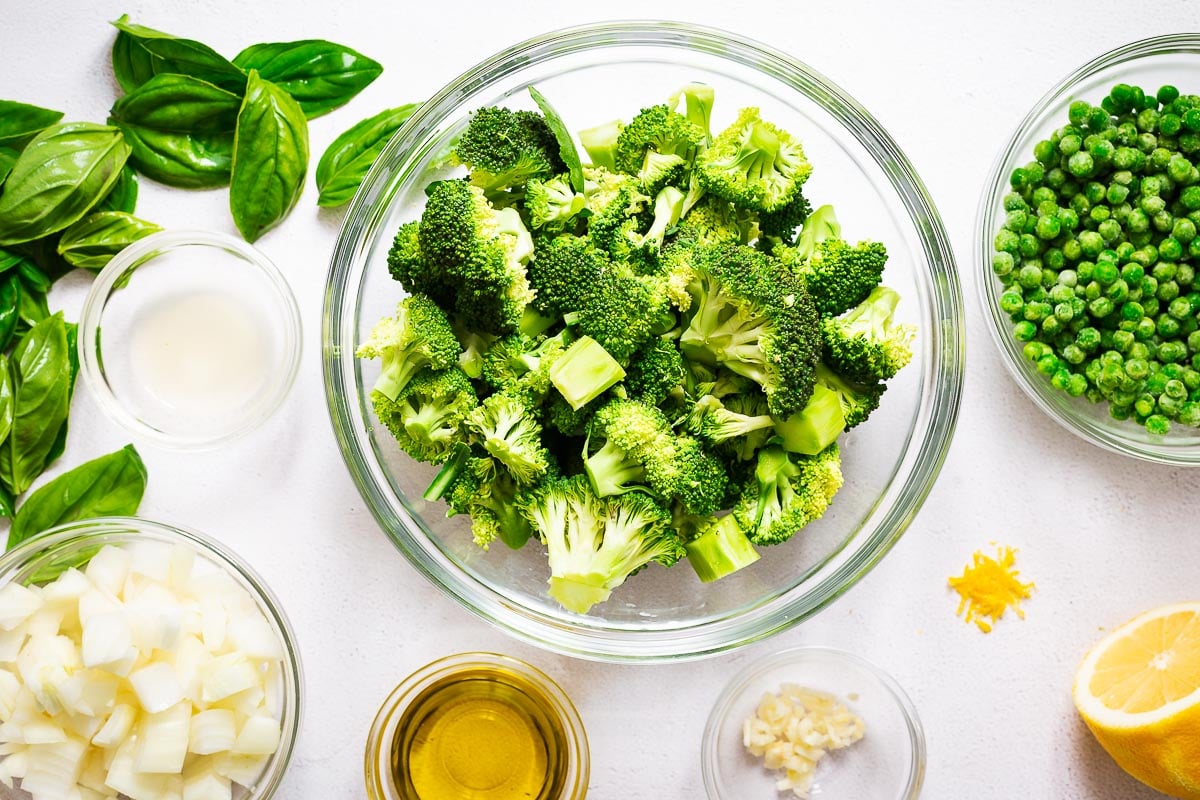 Ingredients for vegan broccoli soup viewed from above. The arrangement include peas, basil, chopped onion, olive oil, garlic and lemon.