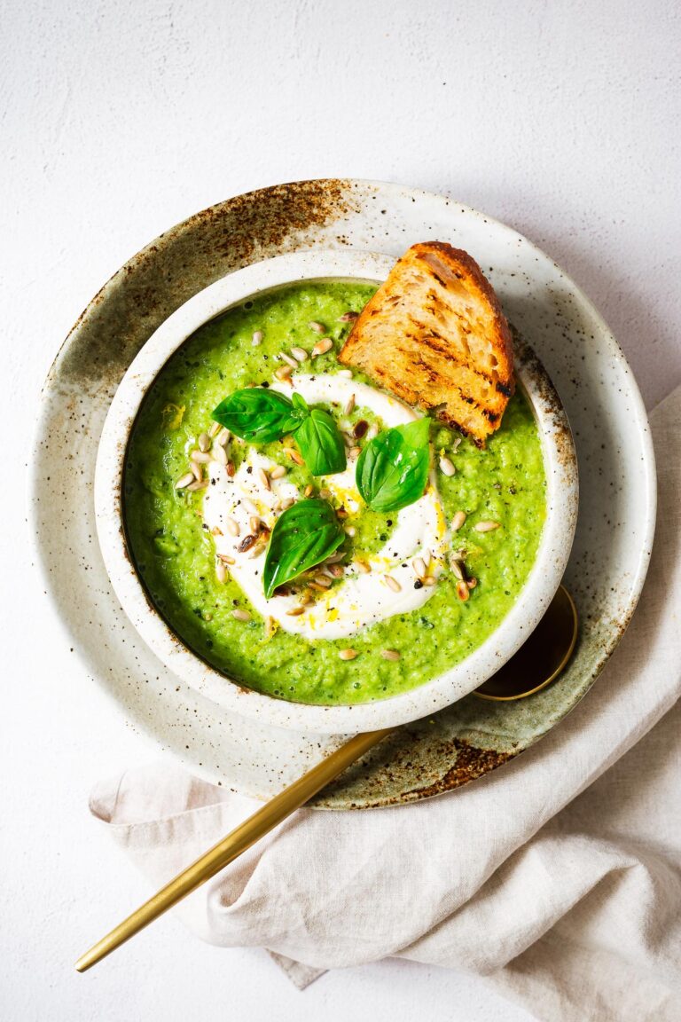A ceramic bowl with vibrant green vegan broccoli soup with cashew cream, sunflower seeds and fresh basil leaves. A slice of sourdough is placed in the soup and there is a gold soup spoon.