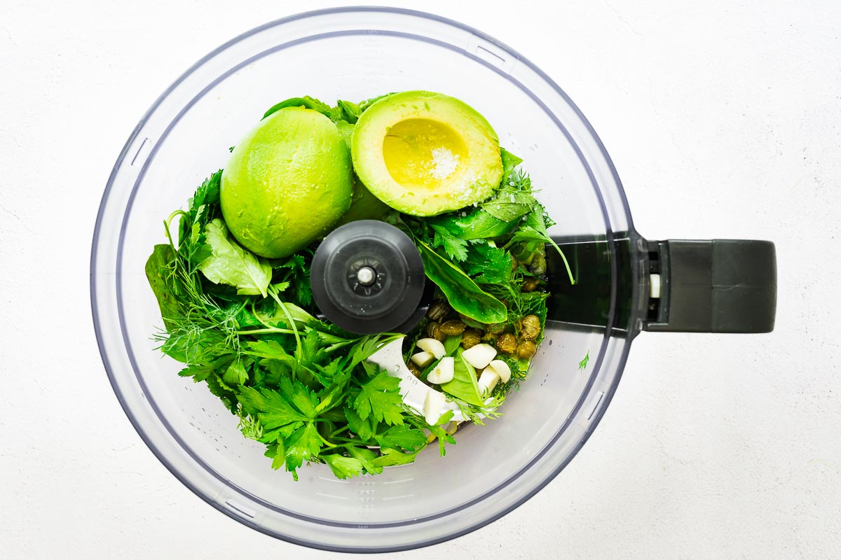 Ingredients for an avocado green goddess sauce in the bowl of a food processor viewed from above.