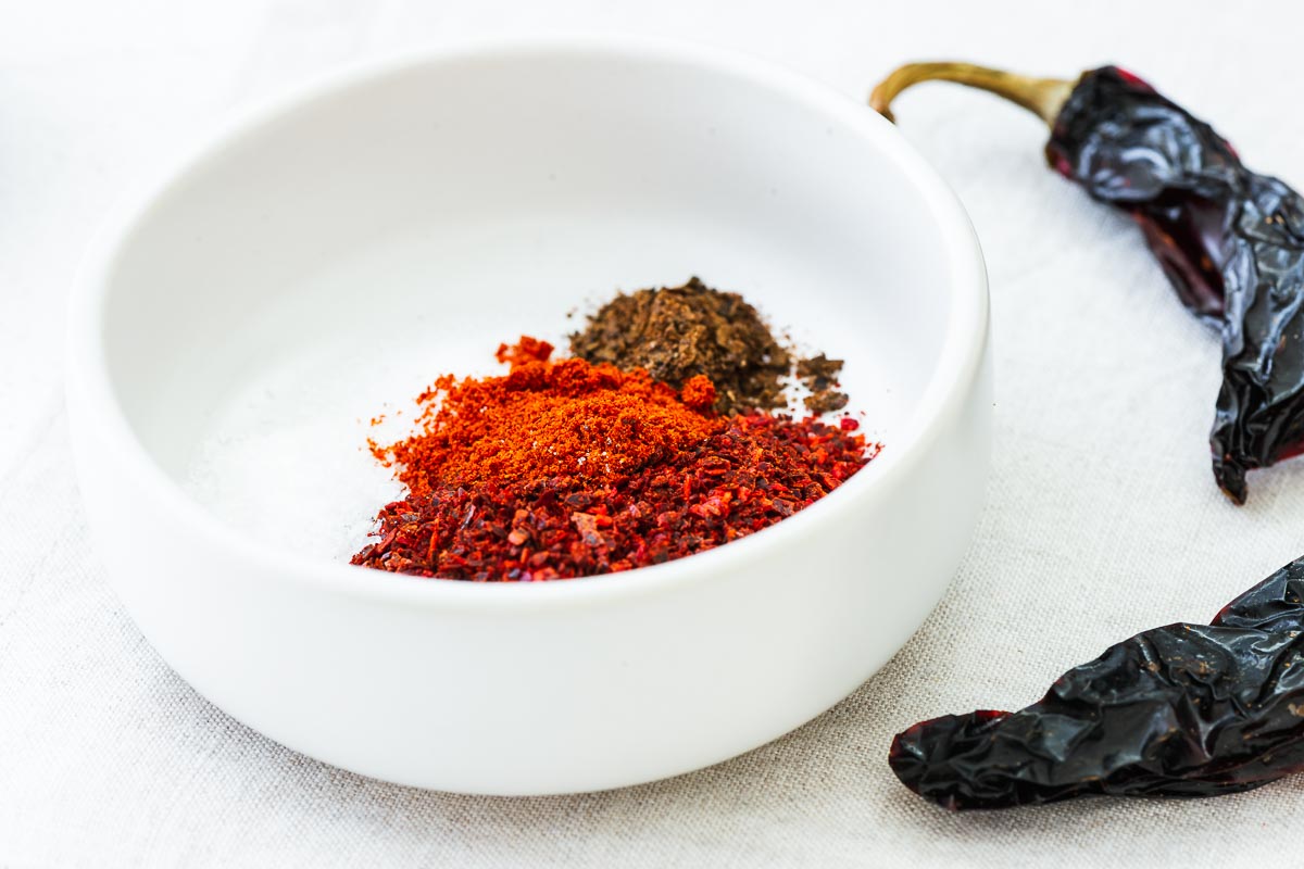 Chilli flakes, smoked paprika, ground black lime, salt and citric acid in a small white bowl next to whole dried chillies.