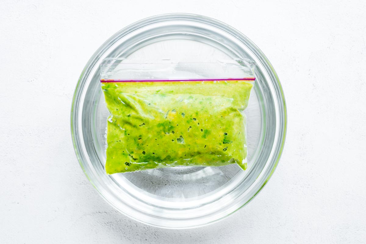 A freezer bag with frozen guacamole defrosting in a bowl of cold water viewed from above.