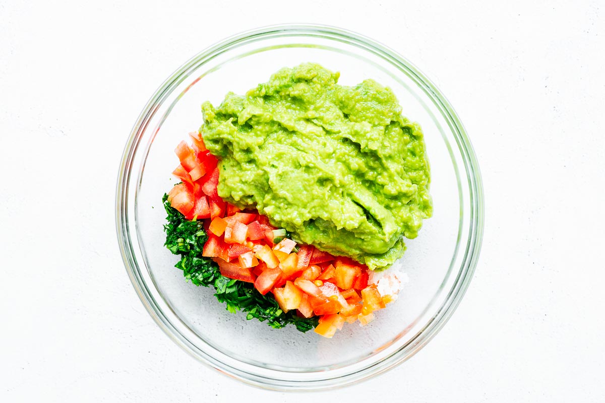 Defrosted mashed avocado with diced tomato, cilantro, onion powder, spring onions and salt. The ingredients are in a glass bowl viewed from above.