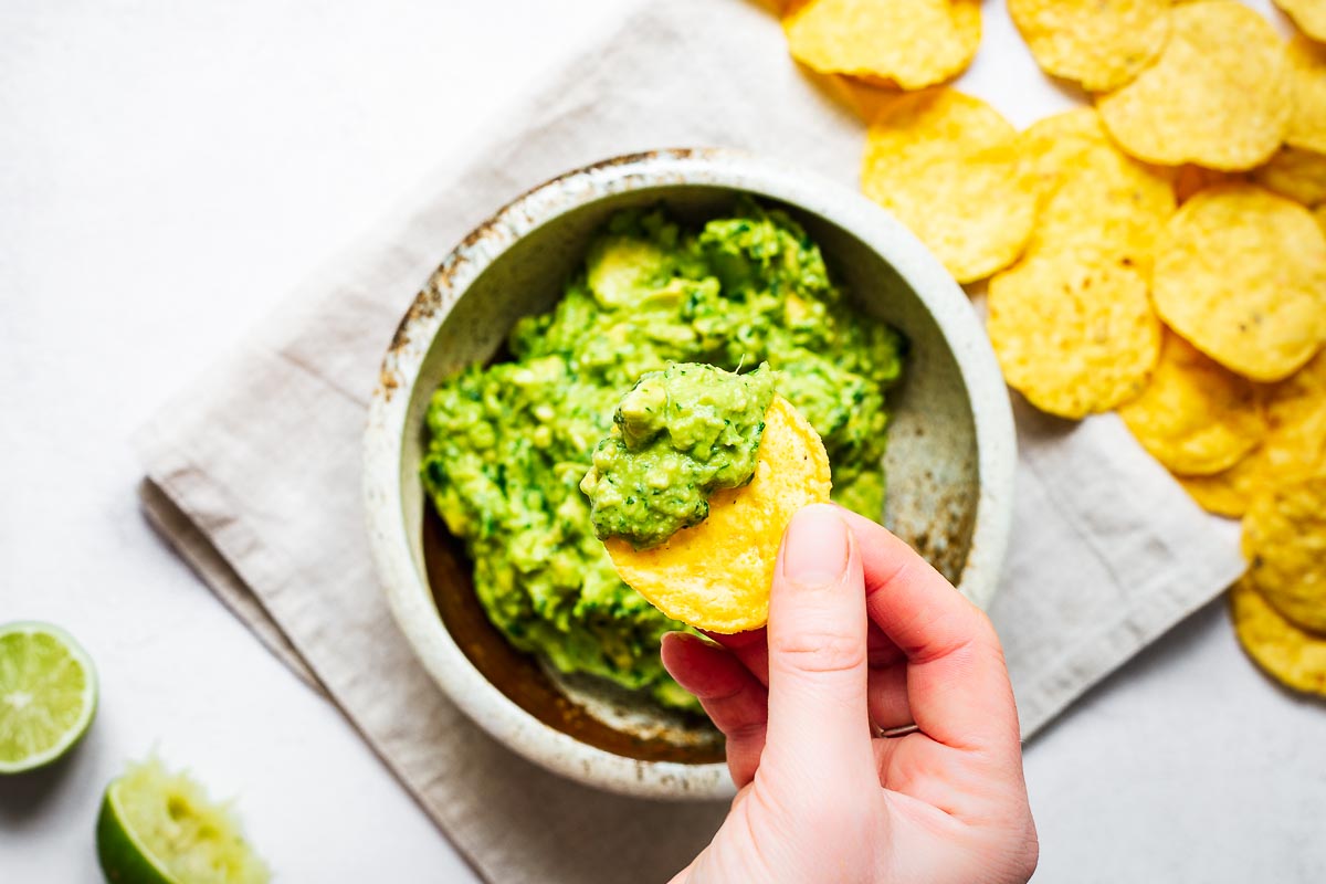 Top down view of a hand using a corn chip to scoop guacamole.