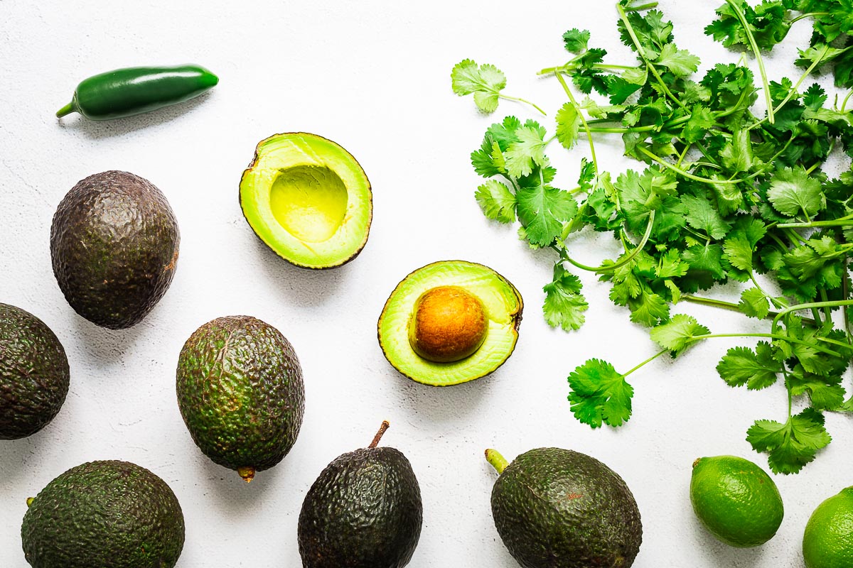 Top down view of guacamole ingredients without tomatoes or onions. It includes sliced avocado, fresh cilantro leaves (coriander leaves), jalapeño and lime.