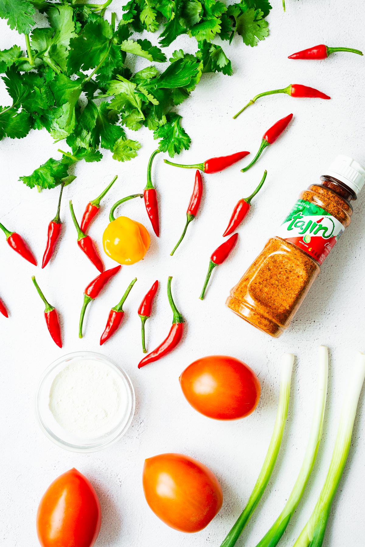Optional flavour boosting ingredients for guacamole, including chillies, tomatoes, green onions, onion powder, cilantro and tajin seasoning. The ingredients are arranged in a flatlay on a light concrete surface.