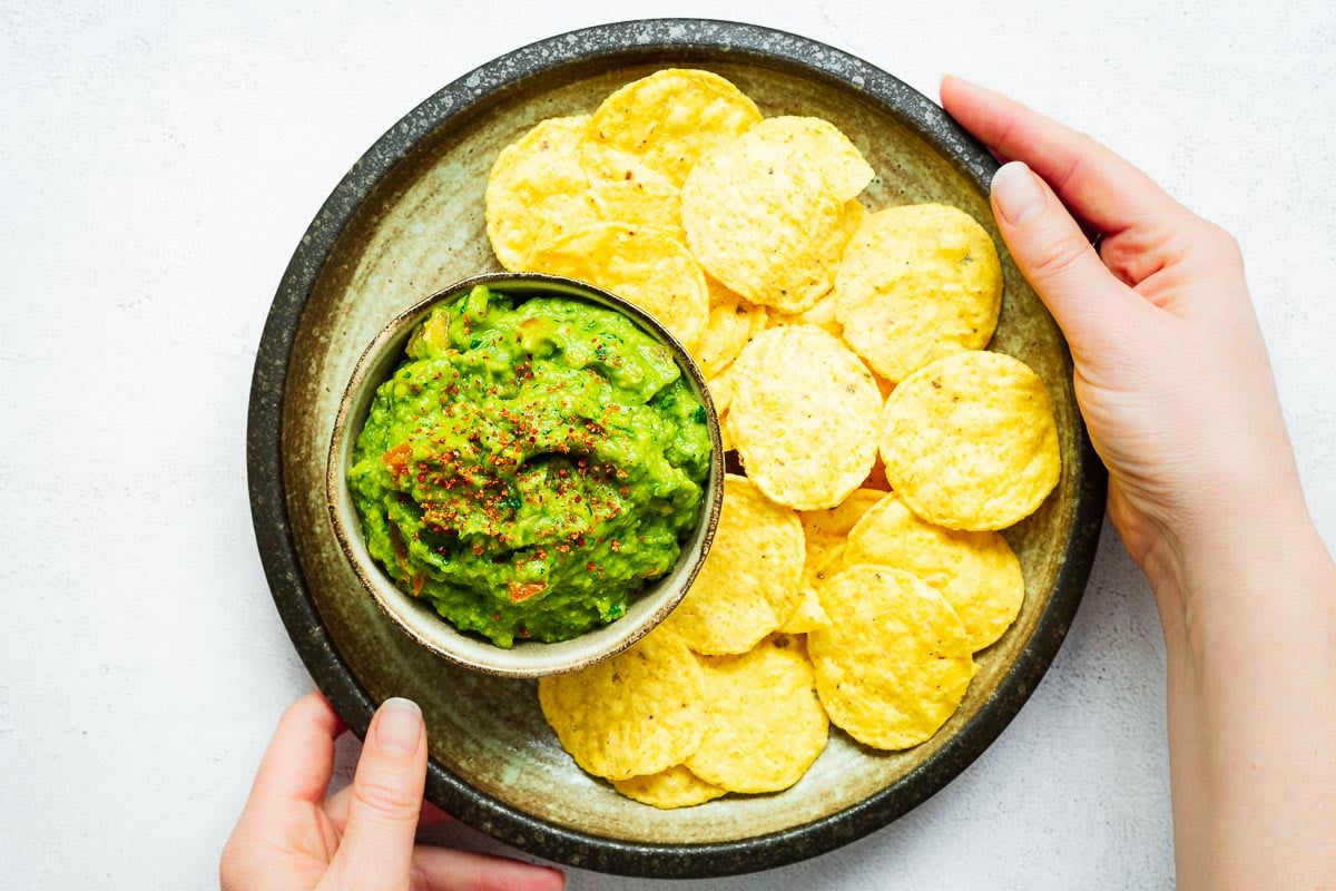 Loaded guacamole in a small bowl surrounded by tortilla chips on a platter being placed down by two hands.