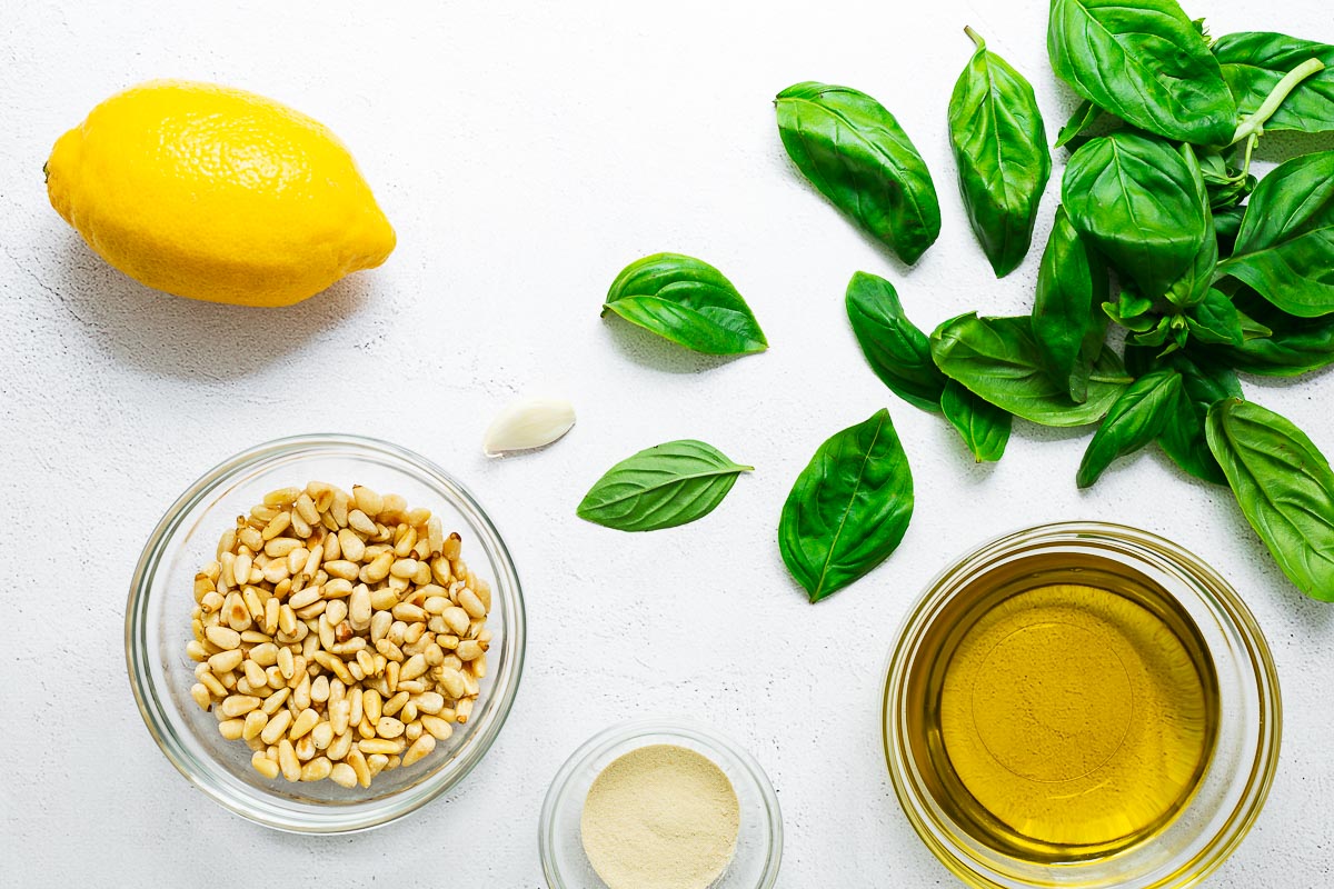 Plant-based basil pesto ingredients arranged in a flatlay, including fresh basil leaves, garlic, pine nuts, lemon, olive oil and nutritional yeast.