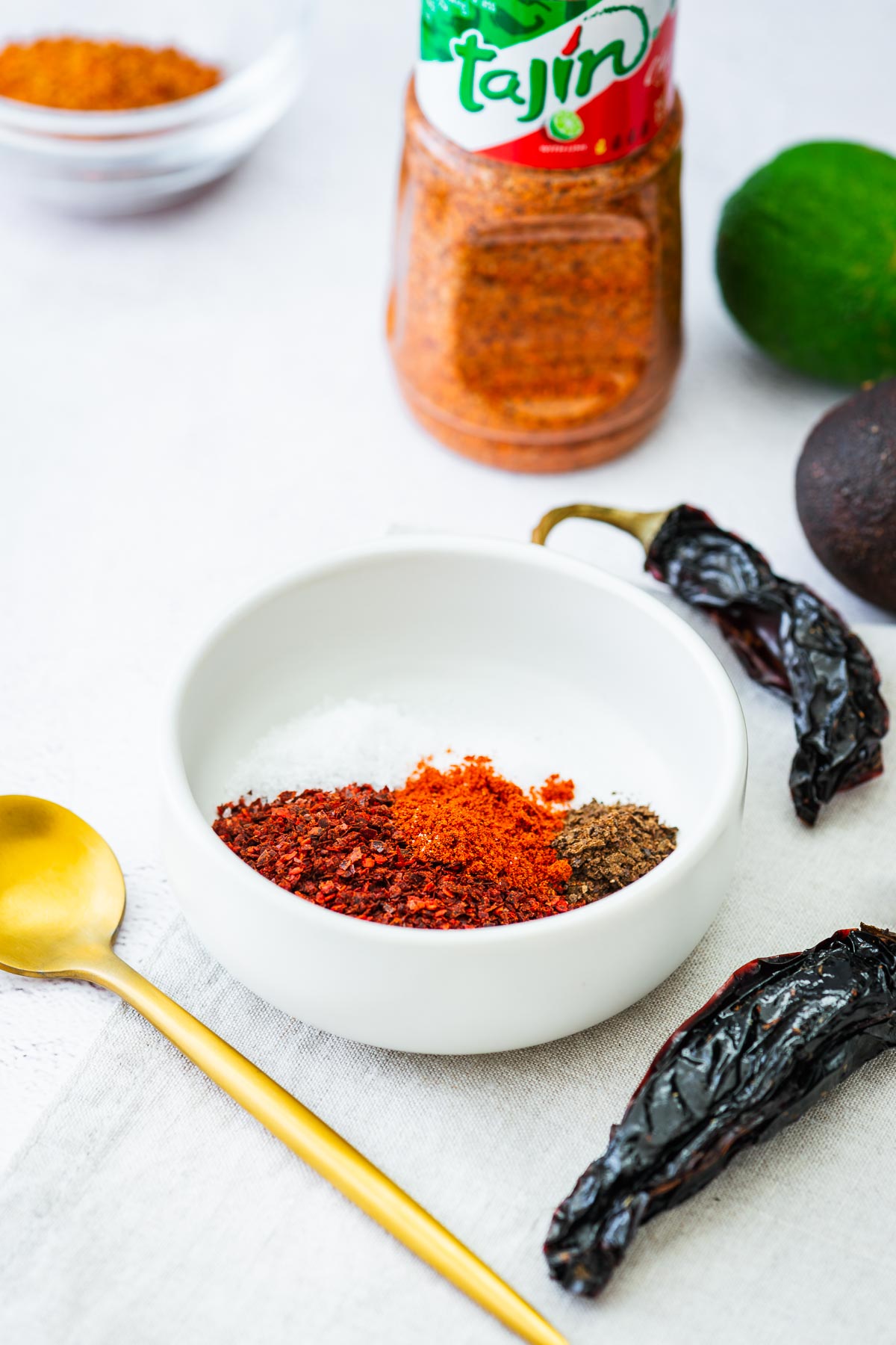 The ingredients for a tajin substitute blend in a small white bowl, including smoked paprika, chilli flakes, black lime powder, salt and citric acid.