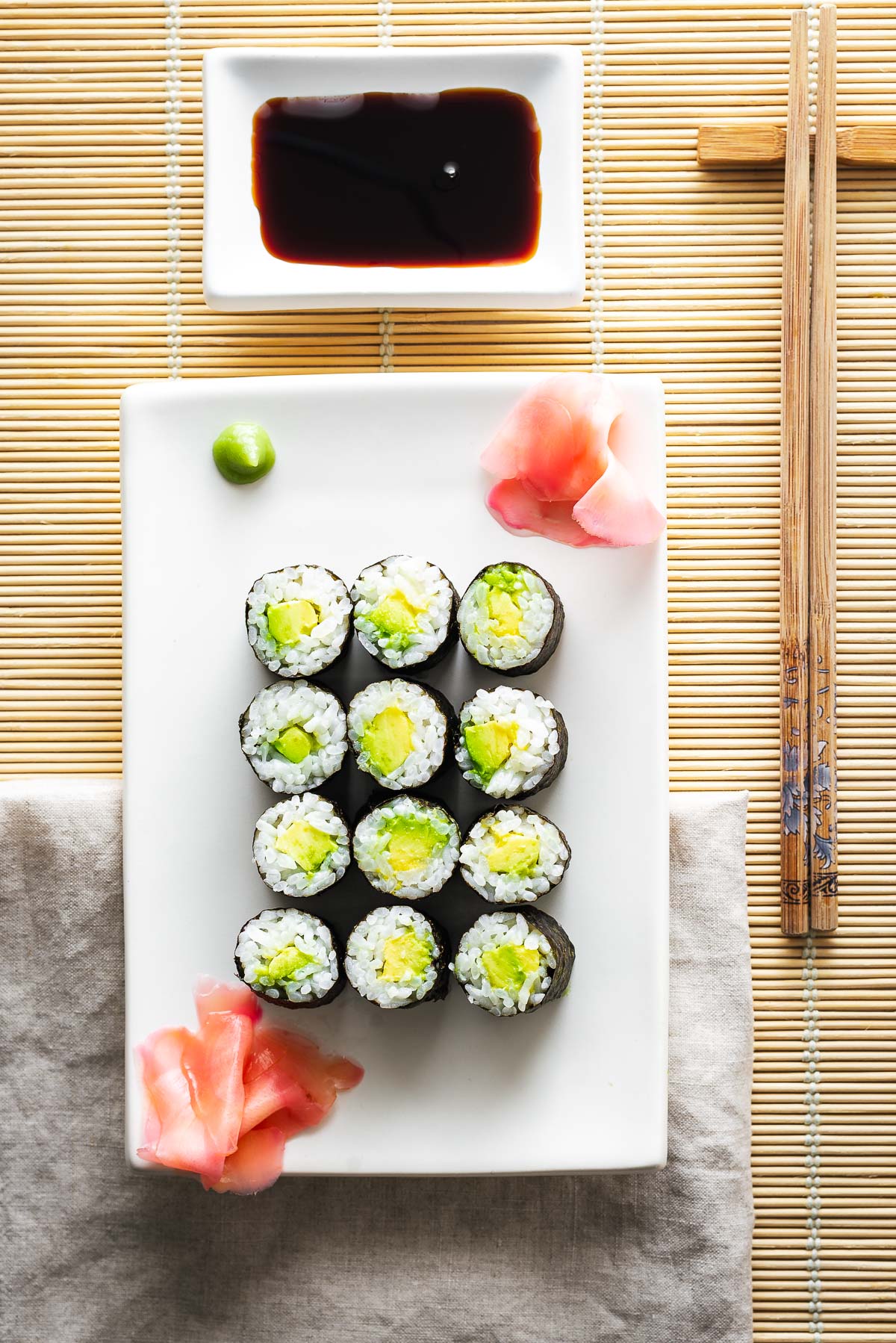 Avocado sushi rolls on a white platter with sushi ginger, wasabi and a bowl of soy sauce with chopsticks.