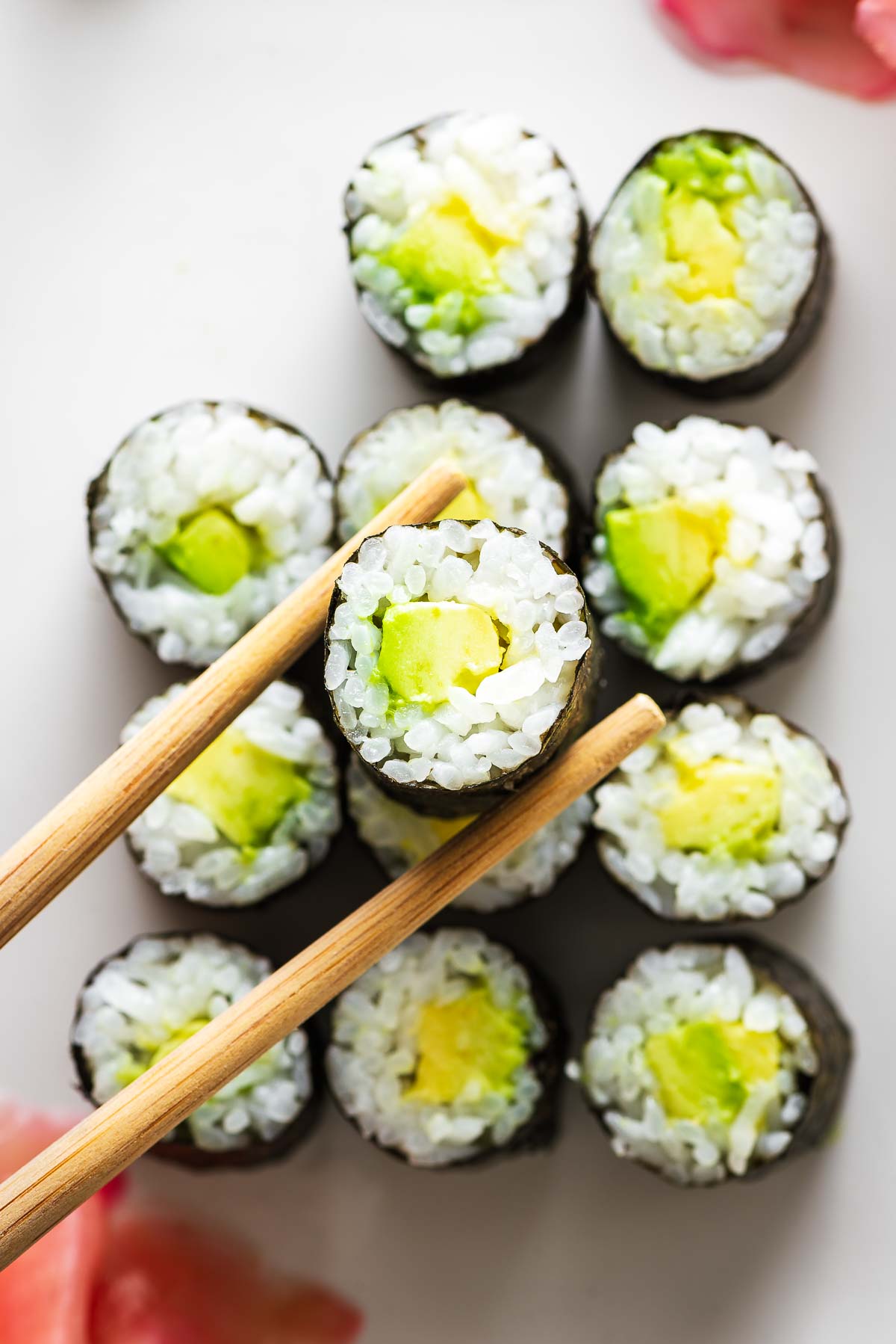 A single avocado sushi roll picked up with chopsticks and more avocado sushi rolls in the background.