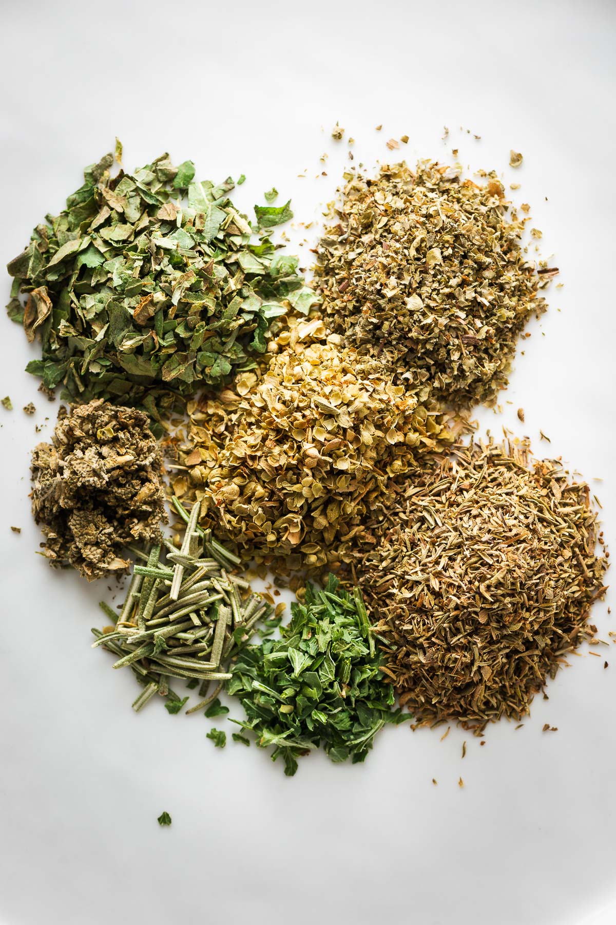 A close-up of small heaps of dried Italian herbs viewed from above.