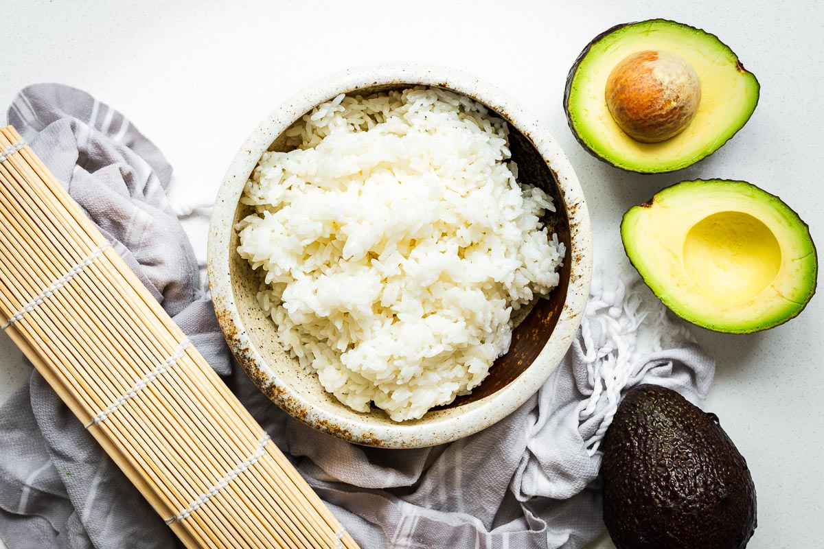 A bowl of white sushi rice, a bamboo placemat, avocados and a damp kitchen towel for homemade sushi.