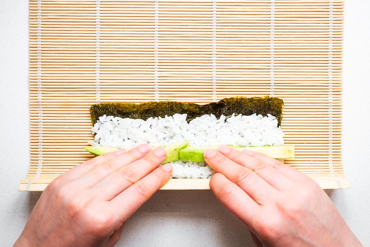 Top down view of nori seaweed with a thin layer of rice and avocado slices being rolled with a bamboo sushi mat.