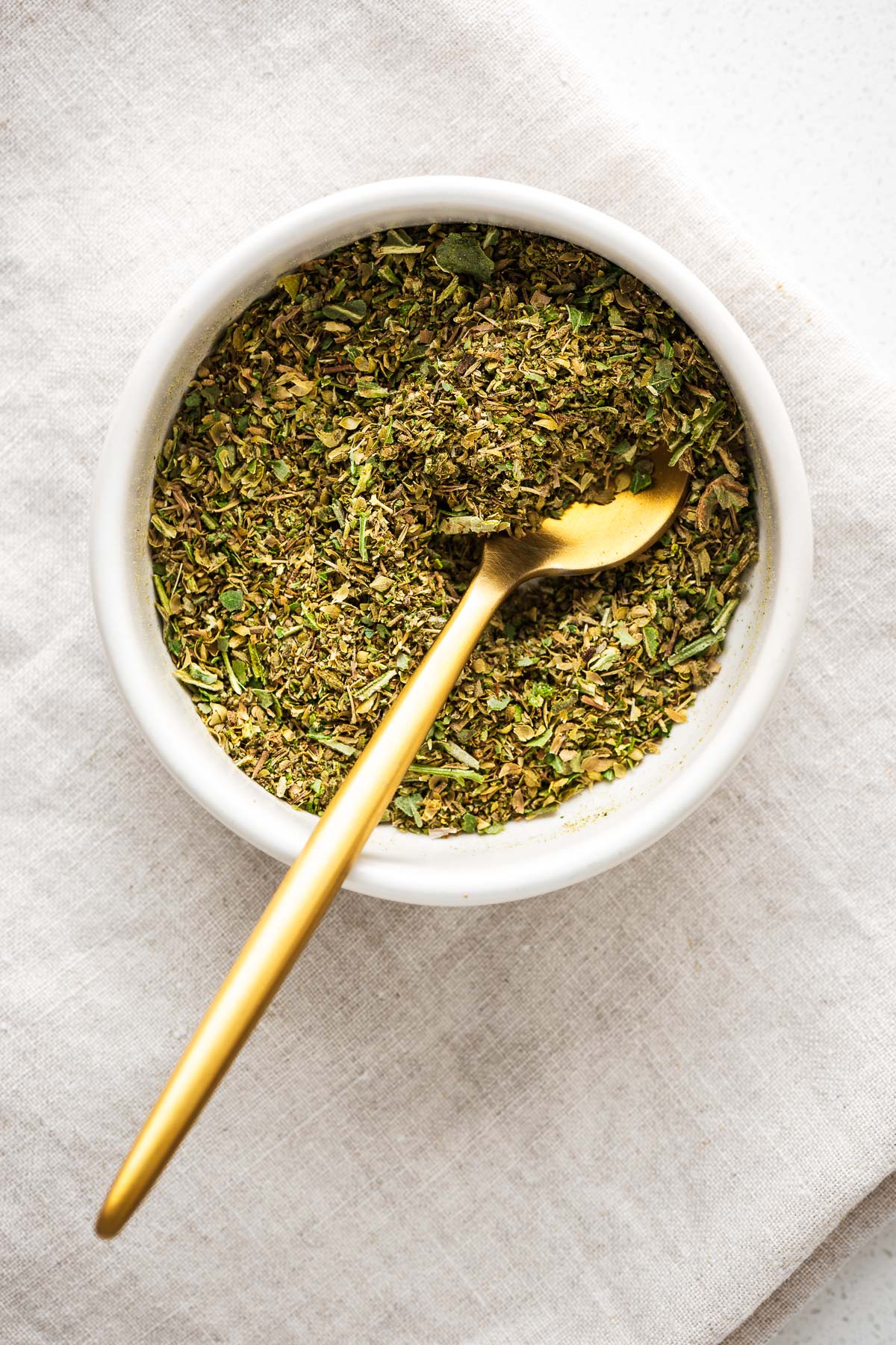 Homemade Italian seasoning in a white bowl with golden teaspoon viewed from above.