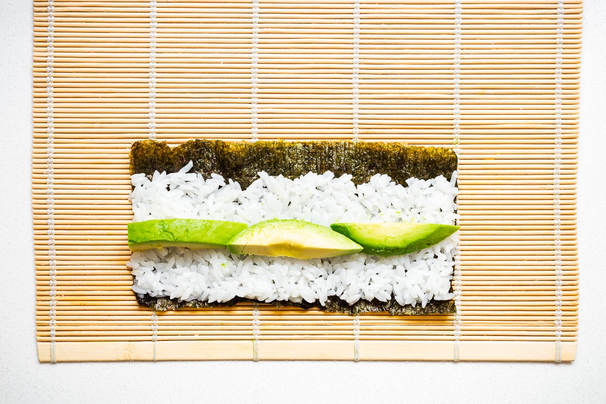 Top down view of nori seaweed with a thin layer of rice and avocado slices on a bamboo mat.