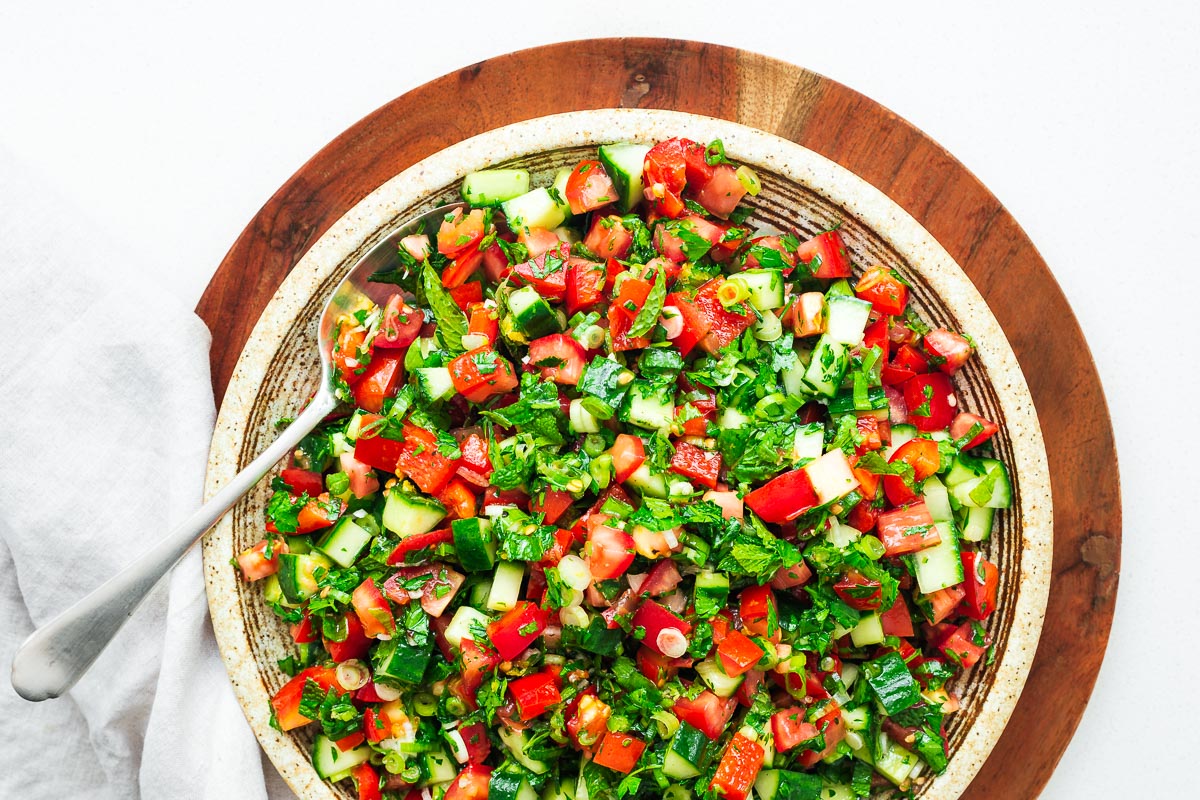Close-up view of a Persian chopped salad in a serving bowl with a serving spoon. The salad is vibrant green and red with tomatoes, red bell pepper, cucumber, fresh mint and parsley and spring onions with other aromatics.