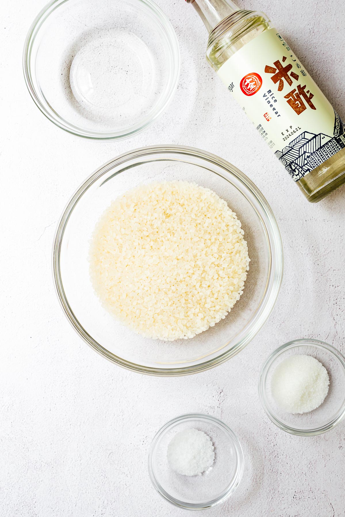 All the ingredients for sushi rice arranged on a concrete surface, viewed from above. It includes a bowl of short-grain rice, rice vinegar, sugar and salt.