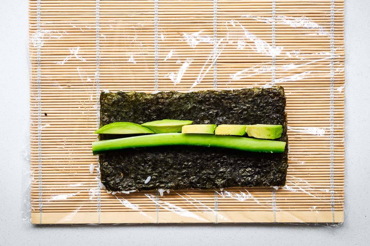 Sliced avocado and cucumber arranged on a nori sheet on a plastic-lined bamboo mat.