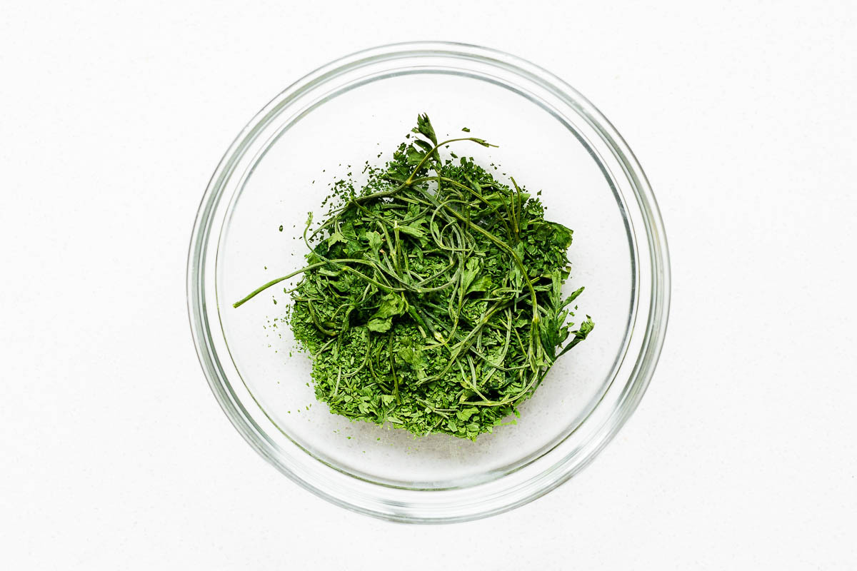 Roughly crumbled oven dried parsley with stems in a glass bowl.